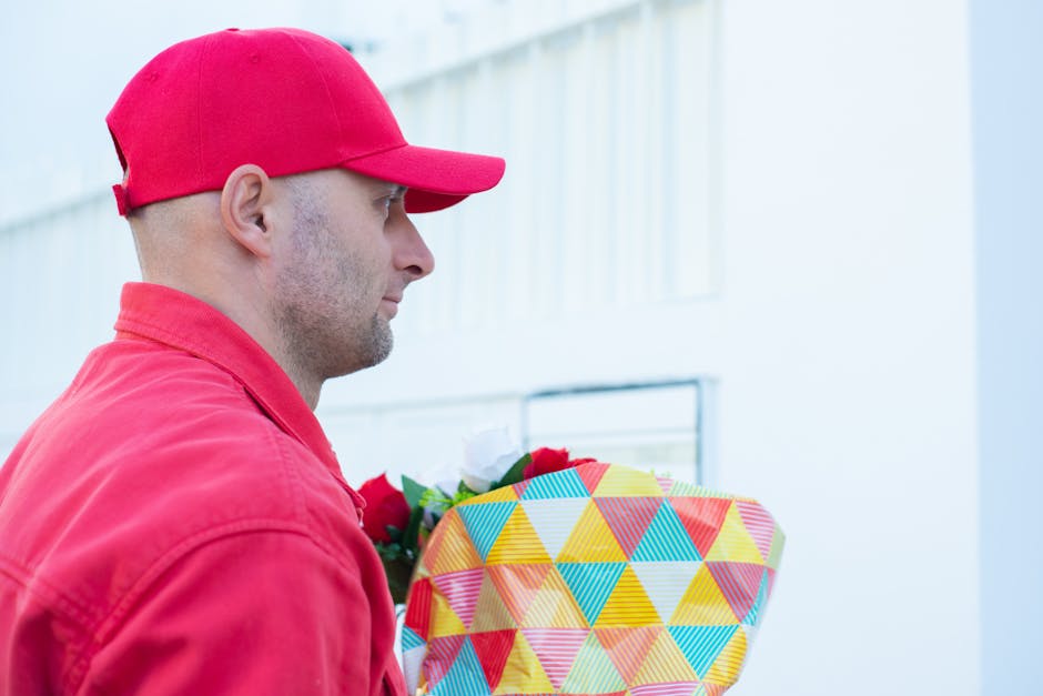 A man dressed in a red cap and red jacket is shown in profile, holding a brightly wrapped floral gift box with a geometric pattern of multicolored triangles. The flowers inside the box are partially v