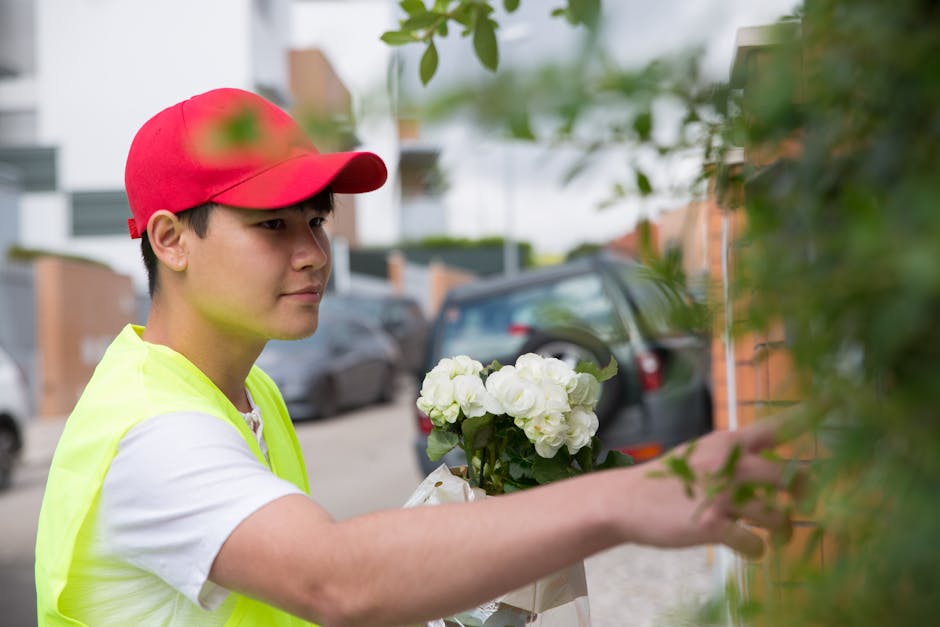 A young male florist wearing a red cap and a high-visibility yellow vest arranging a bouquet of fresh white flowers, likely hydrangeas, with lush green leaves. He is outdoors, standing on a residentia