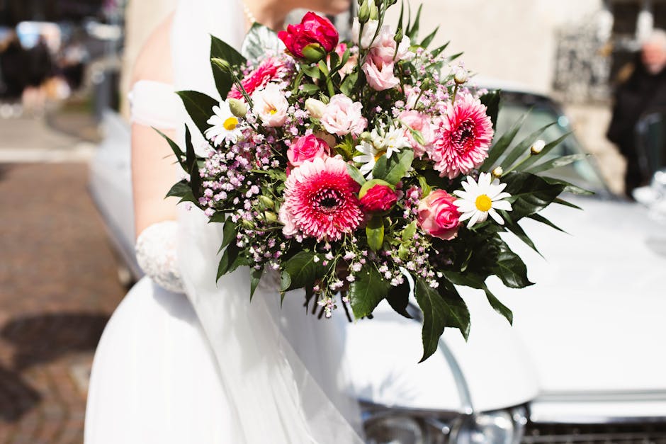 A close-up view of a fresh floral bouquet held by a person dressed in white, featuring pink and white roses, pink gerbera daisies, white daisies with yellow centers, small purple and white filler flow