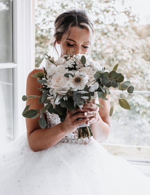 A bride holding a large floral bouquet featuring white roses, white anemones with dark centers, and greenery including eucalyptus leaves. The bouquet is wrapped in a simple, elegant white ribbon. She 