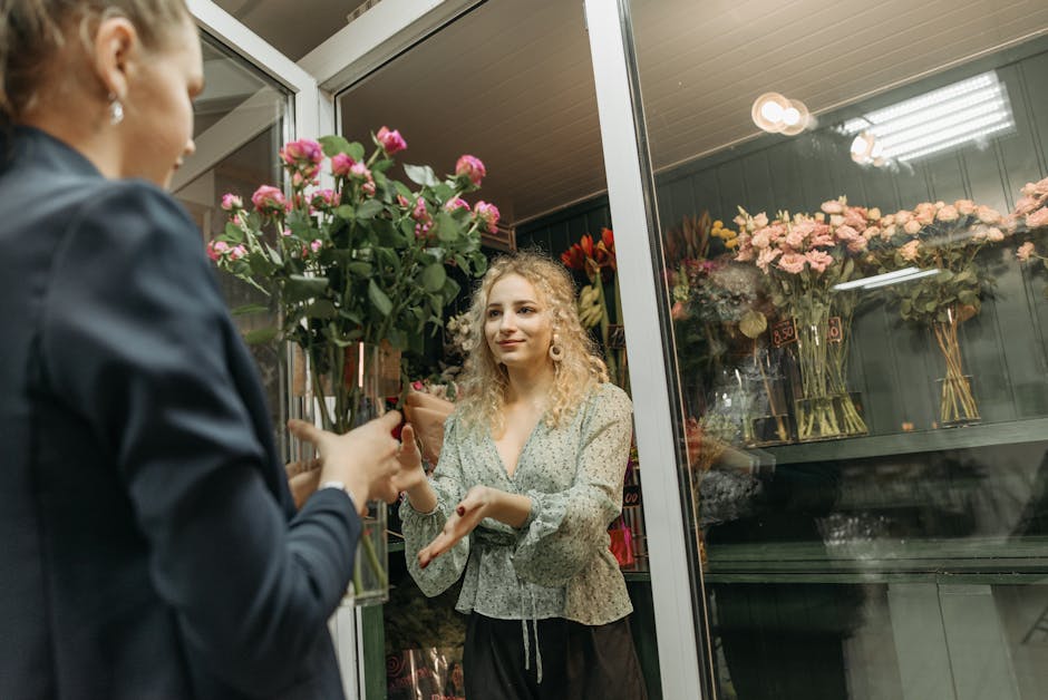 A female florist with curly blonde hair, dressed in a light patterned blouse, is inside a flower shop near Blackheath Station, exchanging a bouquet of pink roses with a customer outside. The bouquet f
