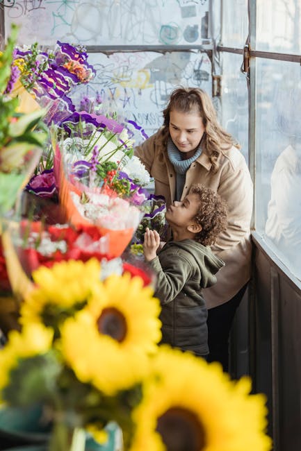 A woman and a young girl are inside a bright, airy flower shop with large windows, examining a vibrant bouquet of mixed flowers. The arrangement includes purple, pink, and red blossoms wrapped in clea